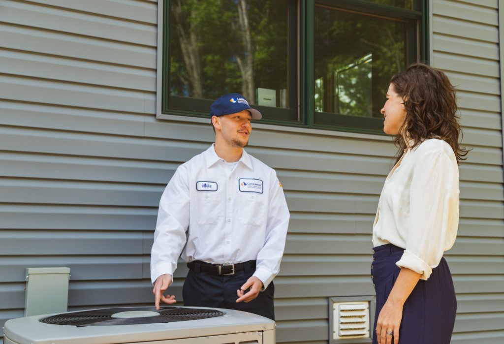 Lancaster Plumbing & Heating employee speaks to homeowner while standing in front of an AC unit outside a gray house